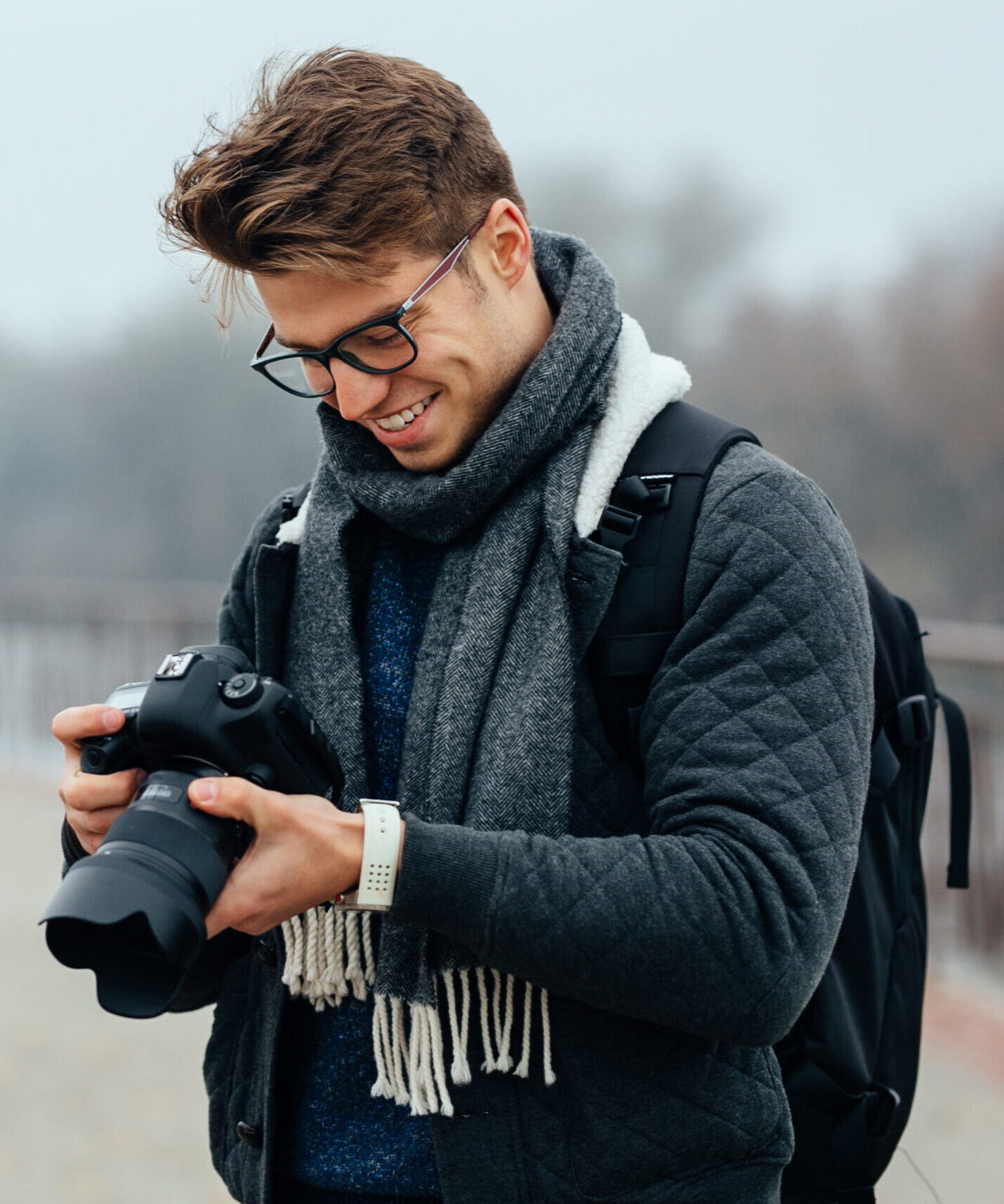 Cheerful handsome man in eyeglasses looks at photos in the camera. Wearing warm stylish jacket, grey scarf, with briefcase. Standing on the street.