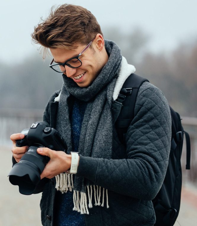 Cheerful handsome man in eyeglasses looks at photos in the camera. Wearing warm stylish jacket, grey scarf, with briefcase. Standing on the street.